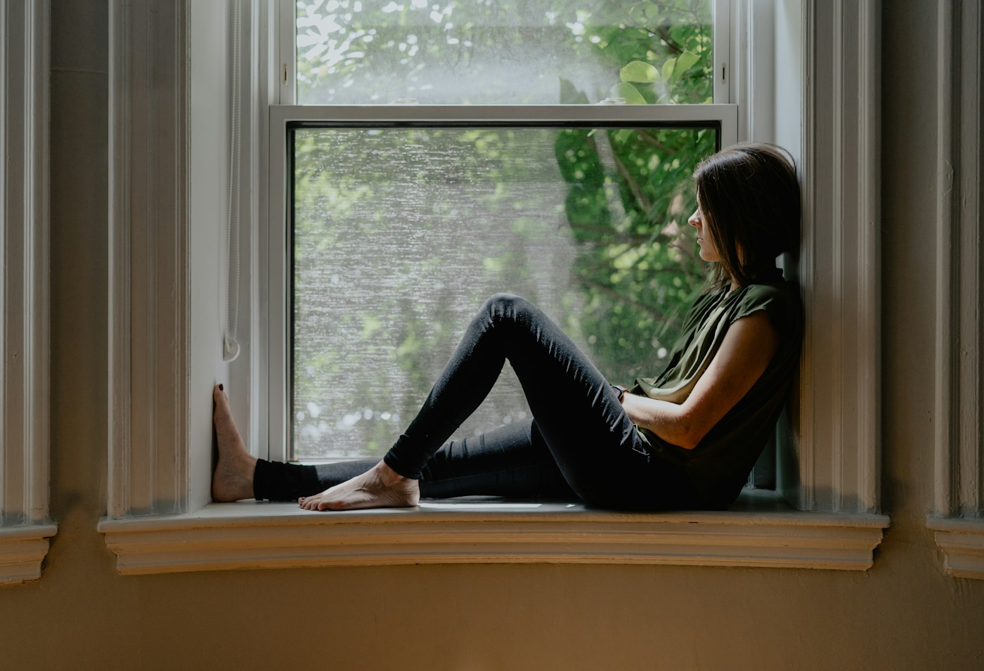A woman sitting on a wide windowsill, looking out of a tall window