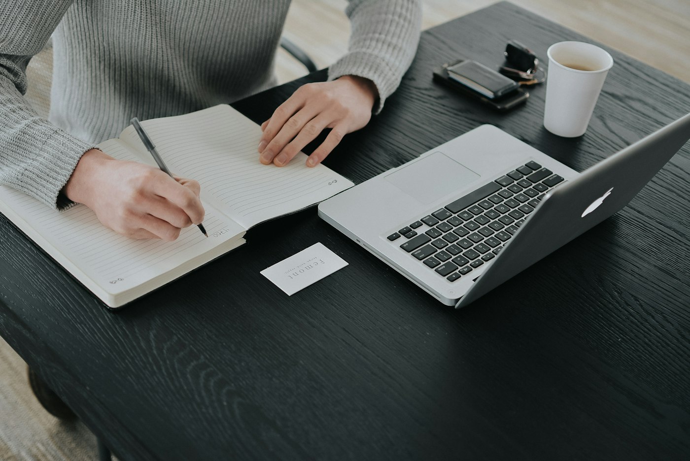 A person writing in a notebook beside an open laptop